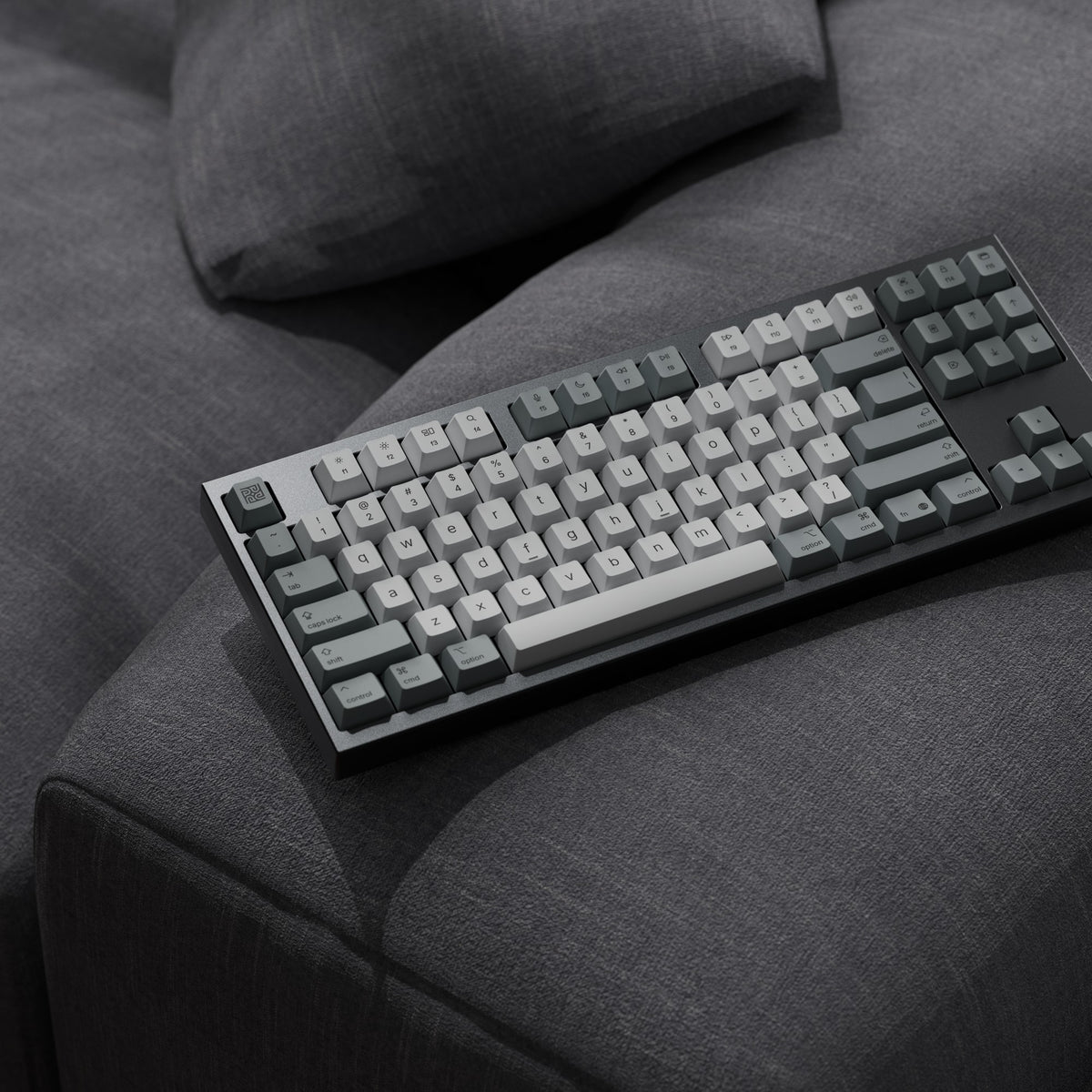 Render: dunkelgraue KBDFans Chamber TKL mit grauen Keycaps auf der Armlehne eines Stoffsofas
